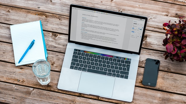 A laptop with a document open on a word processor. A notebook, pen, and glass of water are to the left of the laptop, and a smartphone is to the right.