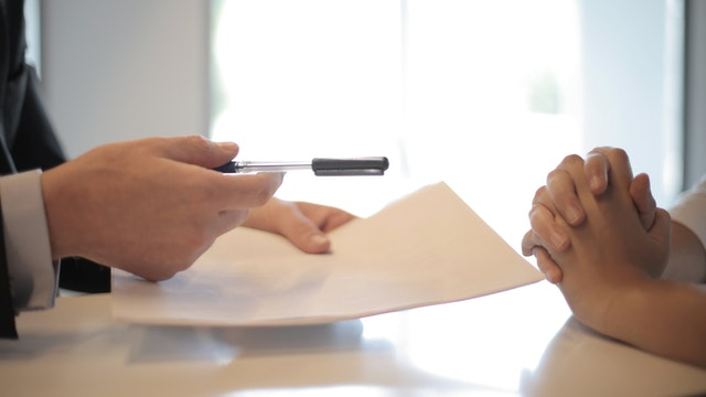A man hands a pile of papers and a pen to a woman with her hands folded.