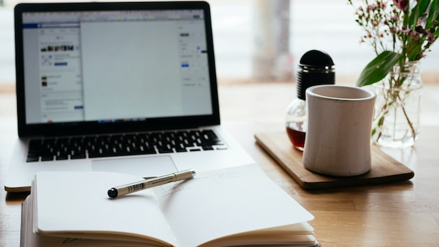 A laptop with a blank doc and a social media site on the screen, surrounded by an open notebook and a coffee mug.