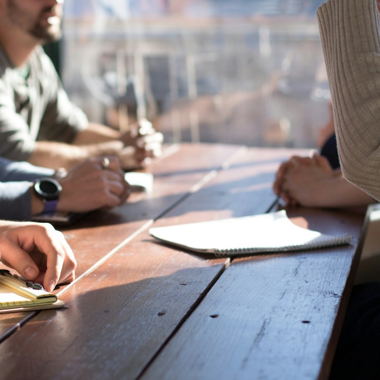 Several people sit around a wooden table with only their hands in the frame. A blank notebook page is in the foreground