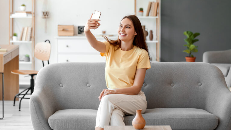 A smiling young brunette sitting on a couch and taking a selfie.