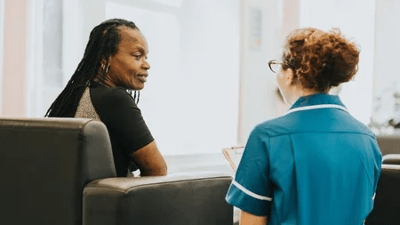 A black woman in an armchair talks to a younger woman with a clipboard.