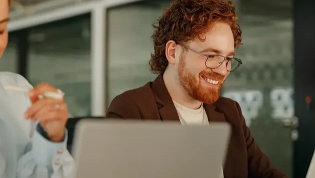 A smiling redhead man working on a laptop.