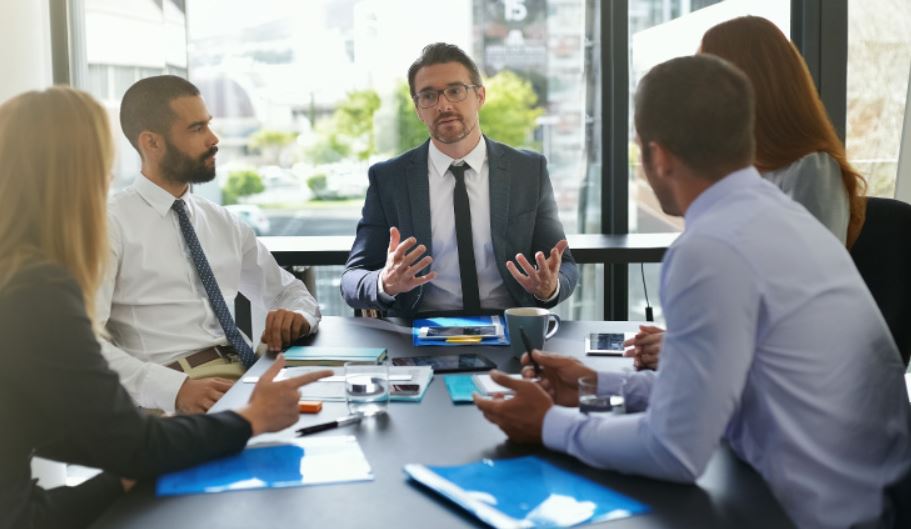 A man sitting at the head of a meeting table talking to four other businesspeople.